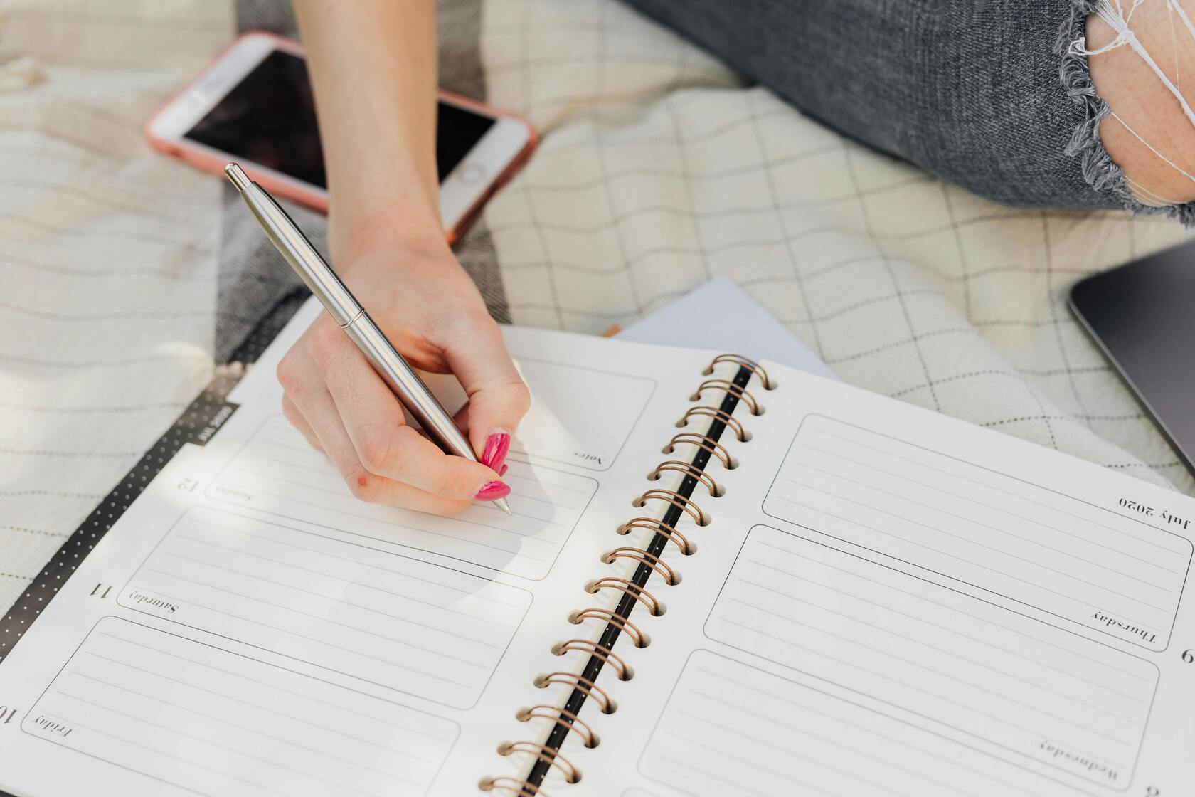 Hand writing in a spiral planner on a bed, with a smartphone and laptop nearby.